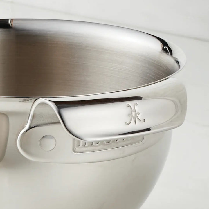 Close-up of a Hestan Provisions stainless steel mixing bowl with handle, featuring a decorative emblem. Part of the 4-piece set, its heat safe to 500°F and dishwasher safe. The shiny surface displays detailed craftsmanship against an off-white background.