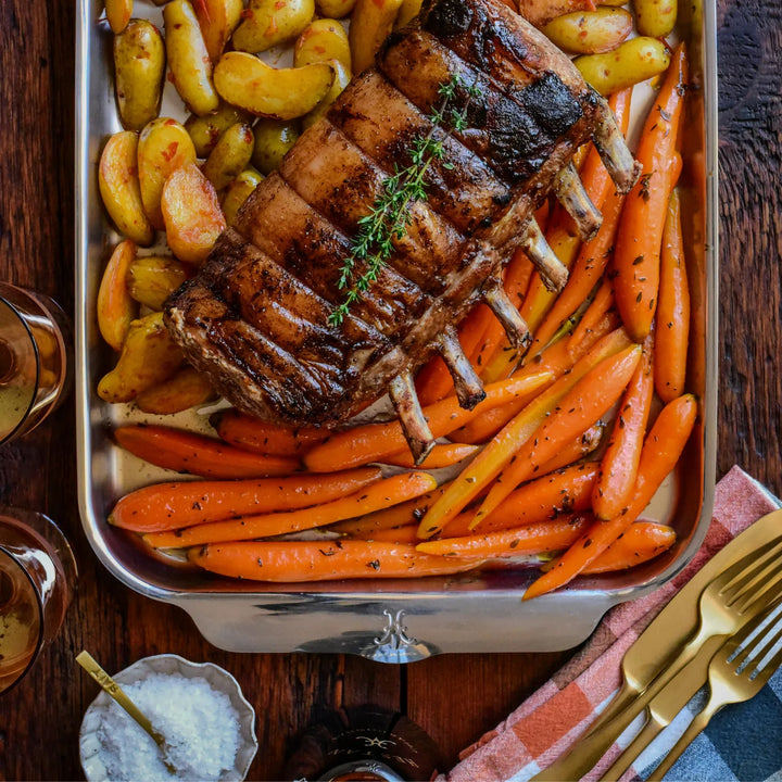 A roasted rack of pork ribs on a Hestan Provisions OvenBond Tri-ply Medium Sheet Pan with Rack, served with roasted baby potatoes and carrots, garnished with herbs. Alongside are two glasses of red wine, a bowl of salt, stainless steel cutlery, and a napkin.