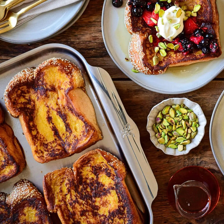 A tray of golden brown French toast sits on a Hestan Provisions OvenBond Tri-ply Medium Sheet Pan with Rack. Nearby, a plated piece is topped with berries, whipped cream, and pistachios, alongside a small bowl of pistachios and a carafe of syrup on the wooden table.