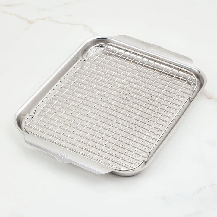 The OvenBond Tri-ply Medium Sheet Pan with Rack by Hestan Provisions is made of stainless steel and sits on a white marble surface. Both the sheet pan and rack are oven-safe, showcasing quality and durability.