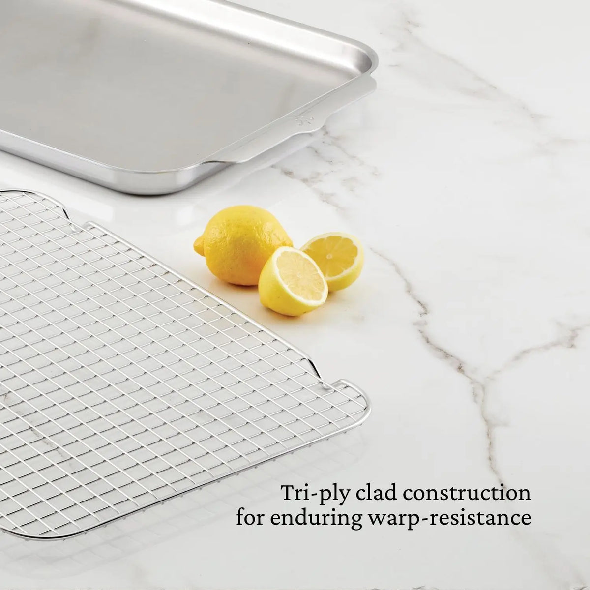 The Hestan OvenBond Tri-Ply Medium Sheet Pan & Rack (12" x 15") sits on a white marble counter with two lemons—one whole, one halved. Text reads: “Tri-ply clad construction for enduring warp-resistance.”.