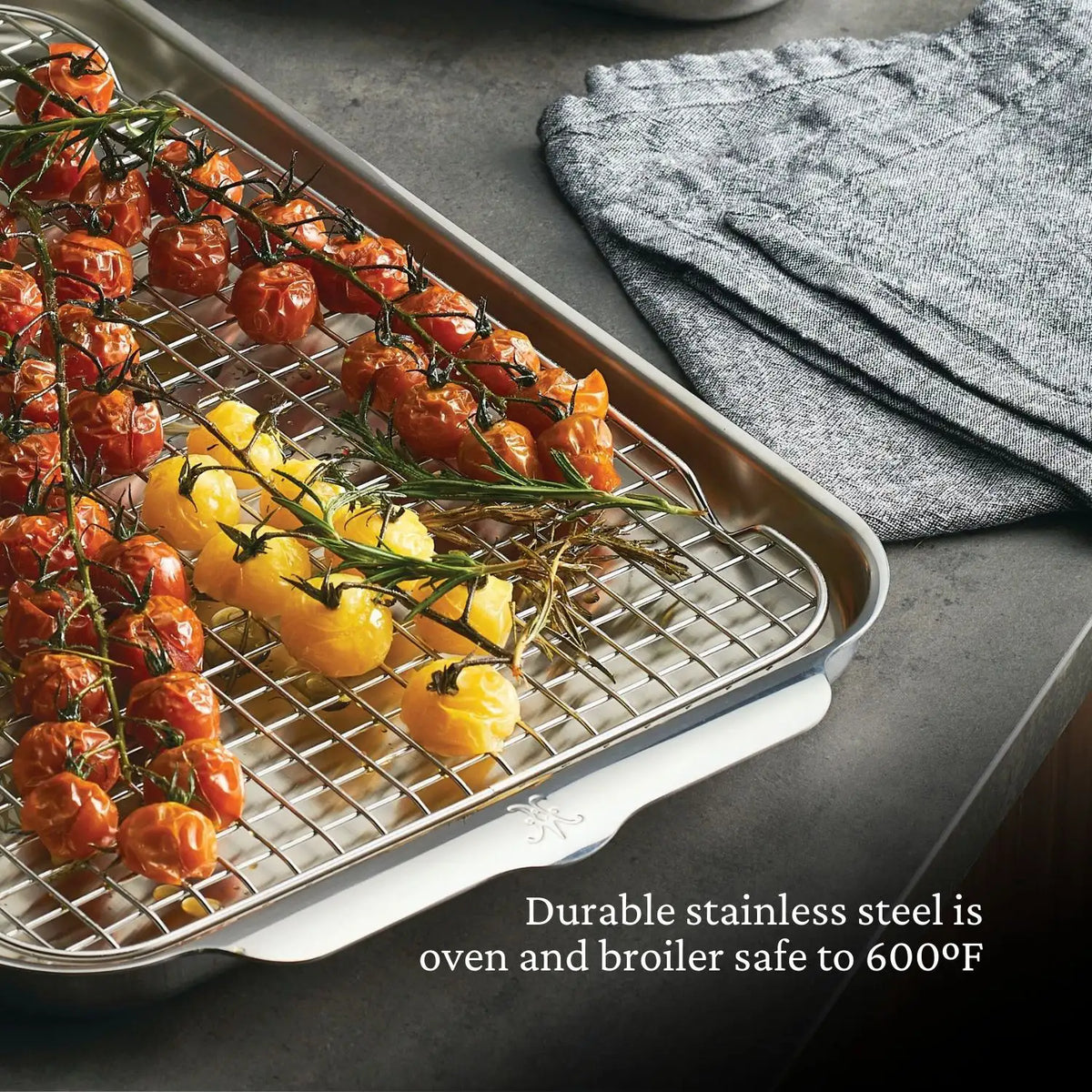 Roasted red and yellow tomatoes rest on a Hestan OvenBond Tri-Ply Medium Sheet Pan & Rack, 12" x 15", beside folded gray towels. Text: “Durable stainless steel is oven and broiler safe to 600°F.”.