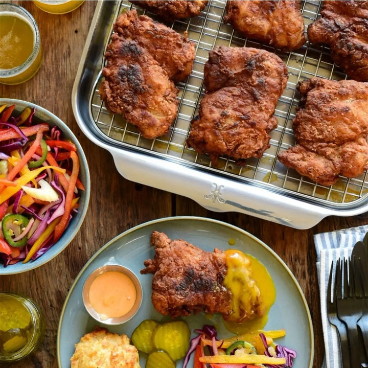 A tray of fried chicken rests on a triple-bonded clad wooden table, while below, the Hestan Provisions OvenBond Tri-ply Medium Sheet Pan with Rack holds fried chicken with pickles, coleslaw, and sauces. Nearby is a bowl of mixed salad alongside drinks and utensils.