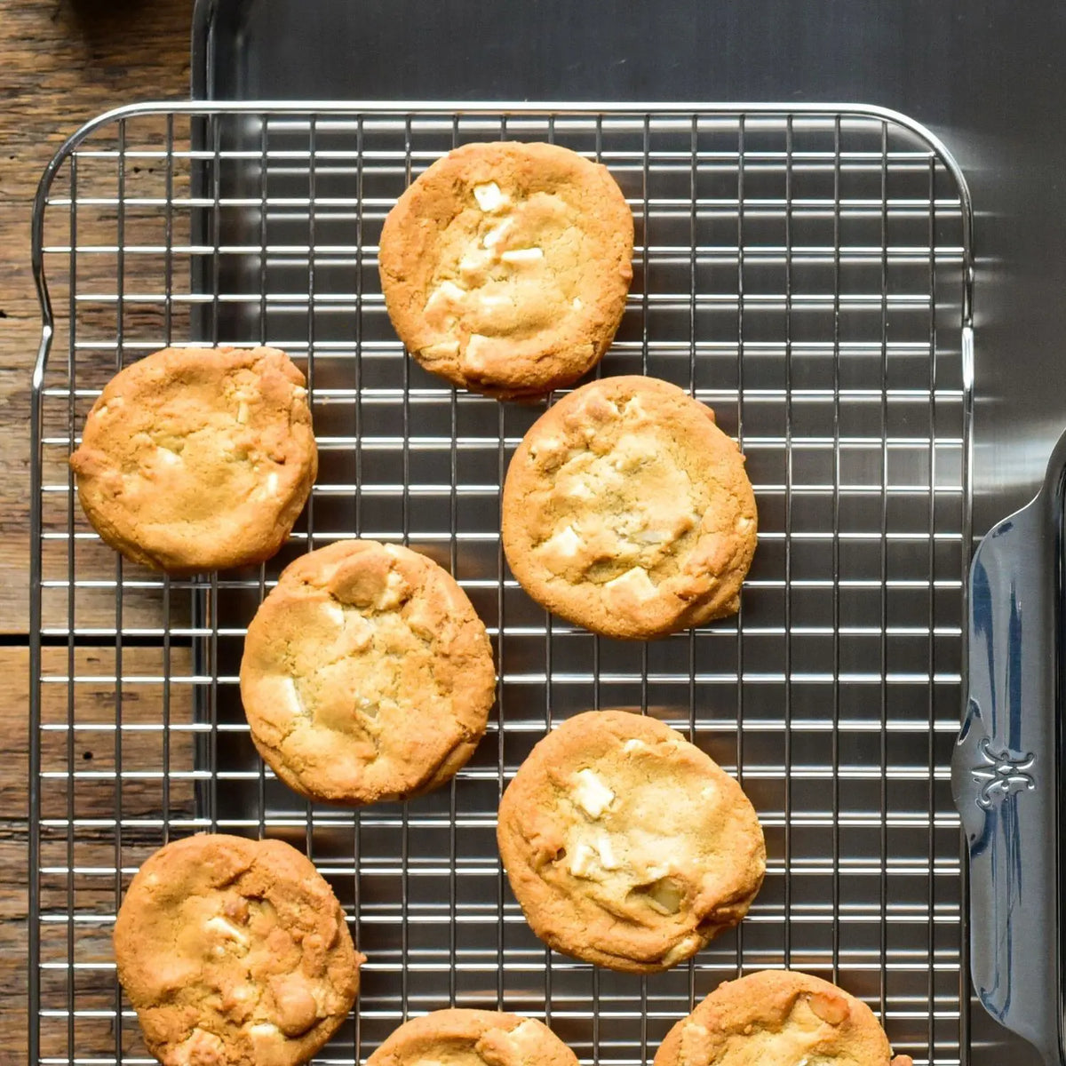Seven round cookies with white chocolate chunks cool on an OvenBond Stainless Steel Medium Pan Rack by Hestan Provisions, placed on a wooden table. A metal spatula is partially visible on the right side of the rack.