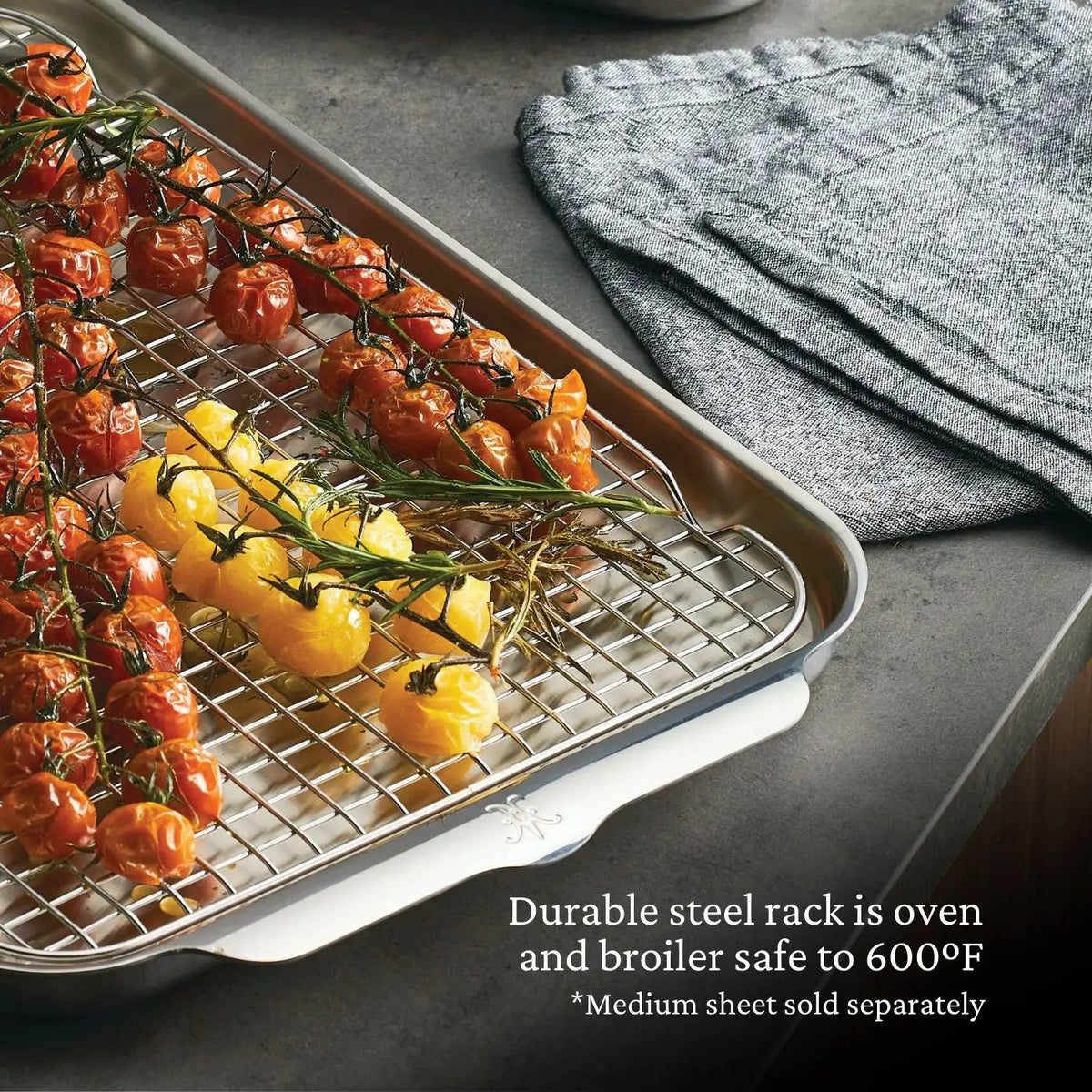 Hestan Provisions OvenBond Stainless Steel Medium Pan Rack roasts cherry tomatoes and small yellow bell peppers on a baking sheet over a gray countertop with folded gray towels nearby. It is oven and broiler safe up to 600°F. *Medium sheet sold separately.