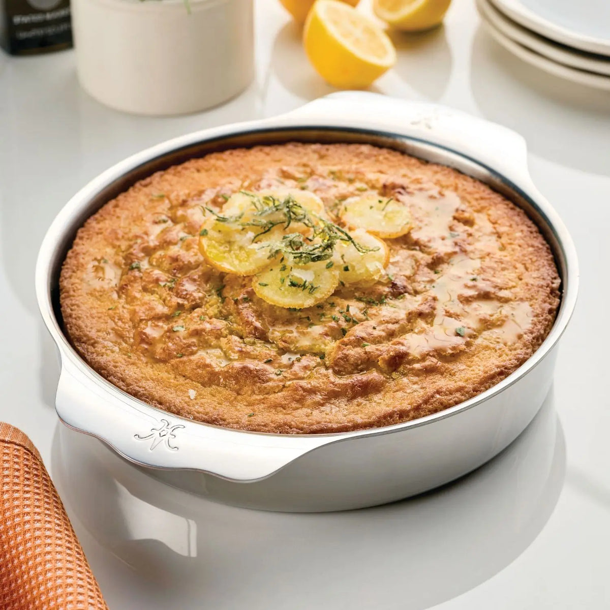 A lemon cake rests gracefully in a Hestan Provisions OvenBond 9-inch Round Baker on a shiny surface. Adorned with glazed lemon slices and herb sprinkles, it exudes freshness. A halved lemon, plates, and an orange kitchen towel add vibrancy to the background.