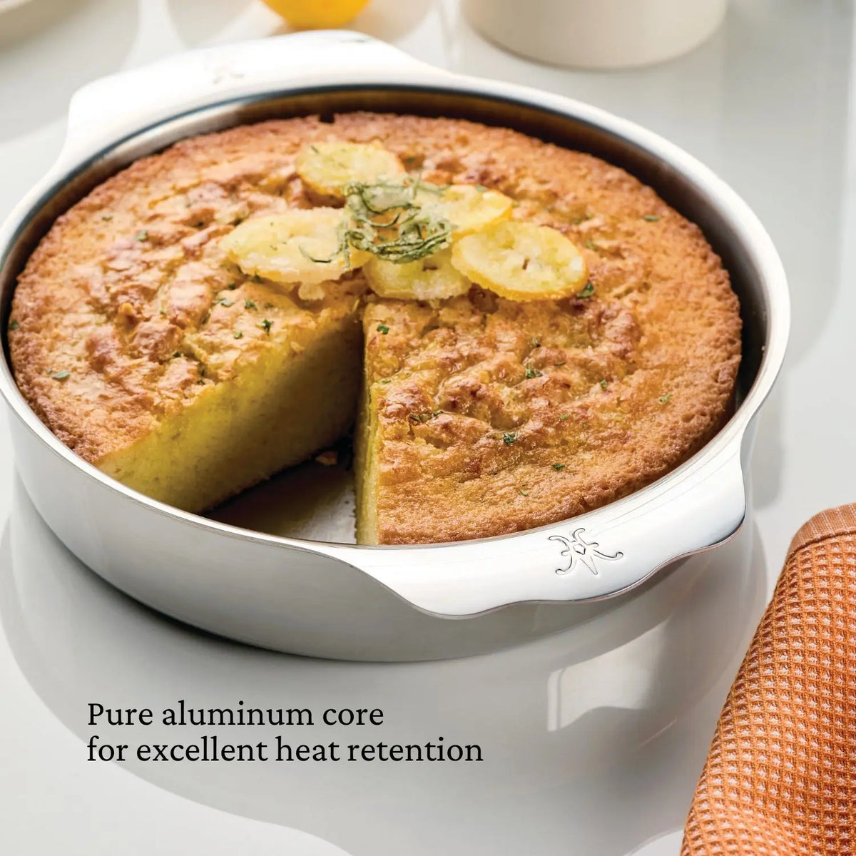 A round cake in a metal baking pan with a missing slice is topped with lemon slices and herbs. Text reads OvenBond Round Baker by Hestan Provisions ensures excellent heat retention. An orange cloth partway visible at the bottom right highlights its broiler-safe, 9-inch design.