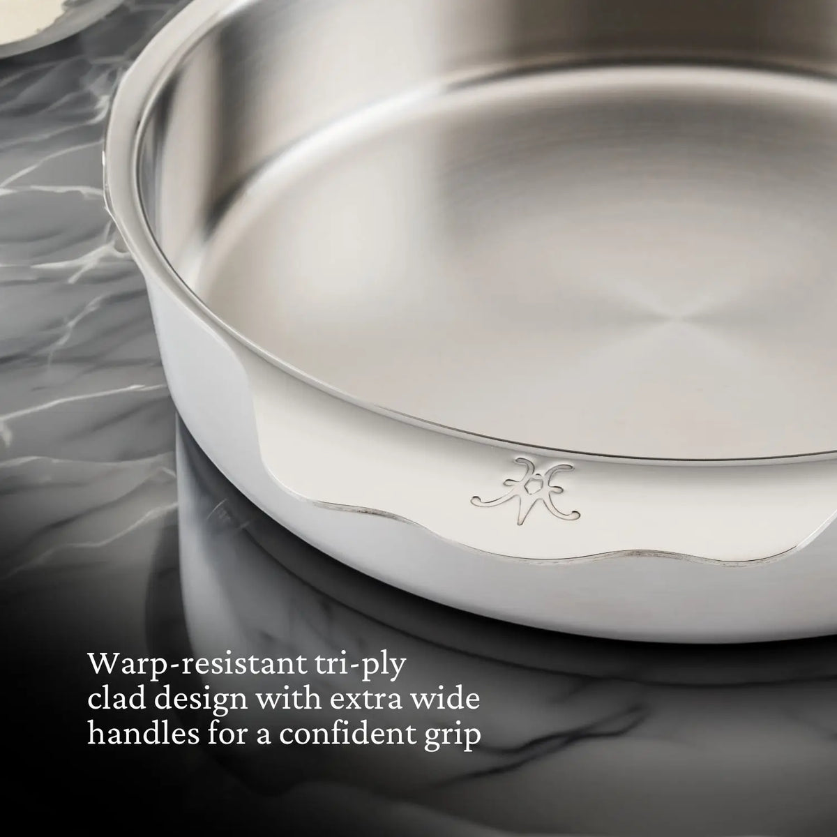 A close-up of the Hestan Provisions OvenBond Round Baker, 9-inch, on a marble surface highlights its warp-resistant Tri-Ply design. The extra wide handles ensure a confident grip. Text in the corner details its broiler-safe features.