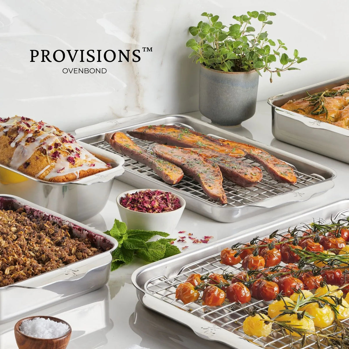 An assortment of stainless steel baking trays and an OvenBond Round Baker, 9-inch, rest on the counter displaying roasted cherry tomatoes, glazed loaf cake, granola, sausages with vegetables, and a potted herb. The word Hestan Provisions is visible in the background.