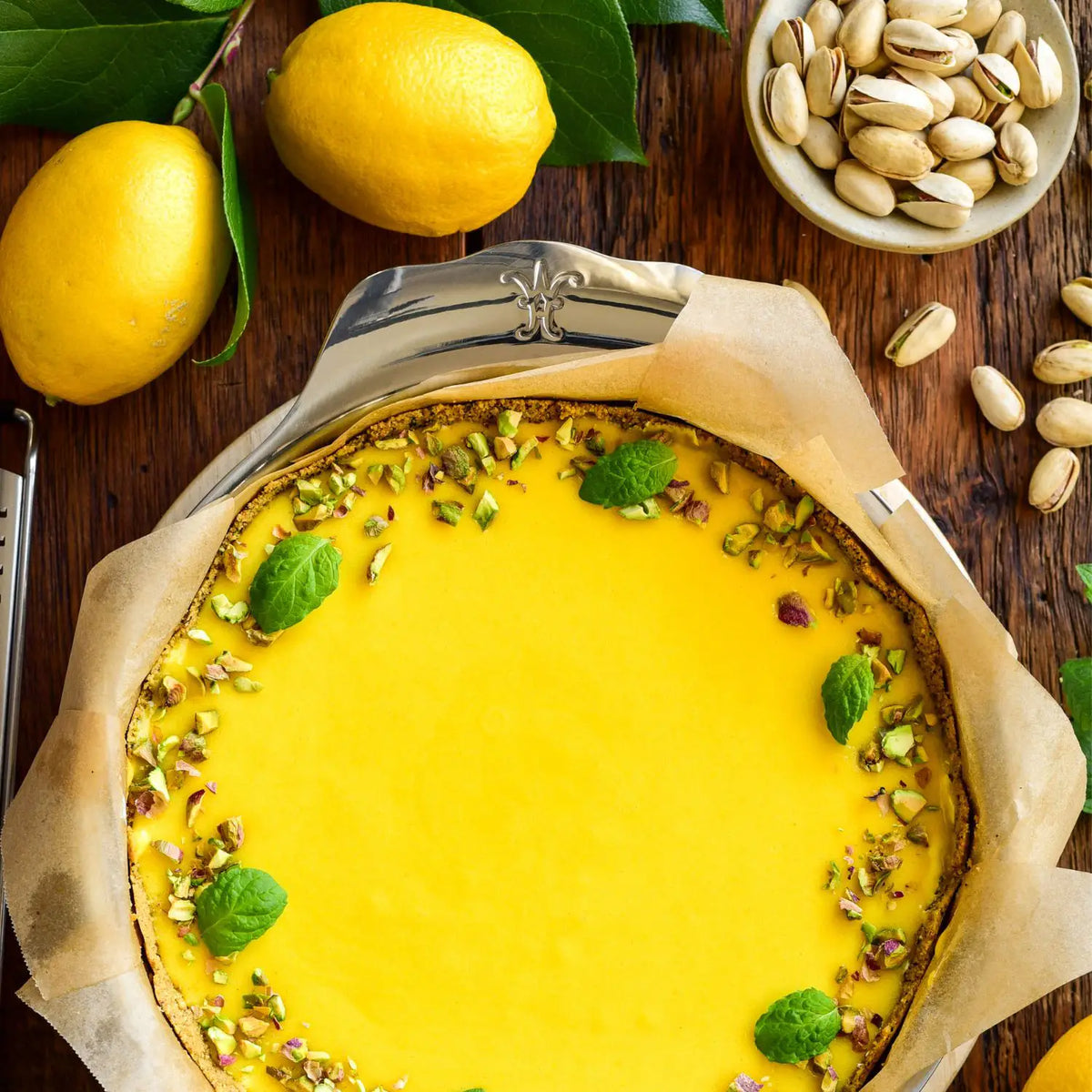 A lemon tart with a yellow filling, topped with pistachios and mint, is baked in the Hestan Provisions OvenBond 9-inch Round Baker, and sits on parchment paper surrounded by whole lemons and pistachios on a wooden surface.