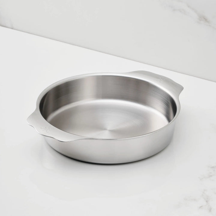 The Hestan Provisions OvenBond 9-inch Round Baker, featuring tri-ply stainless steel and dual handles, sits on a white countertop with a marble-patterned backsplash—oven safe and perfect for all your baking needs.