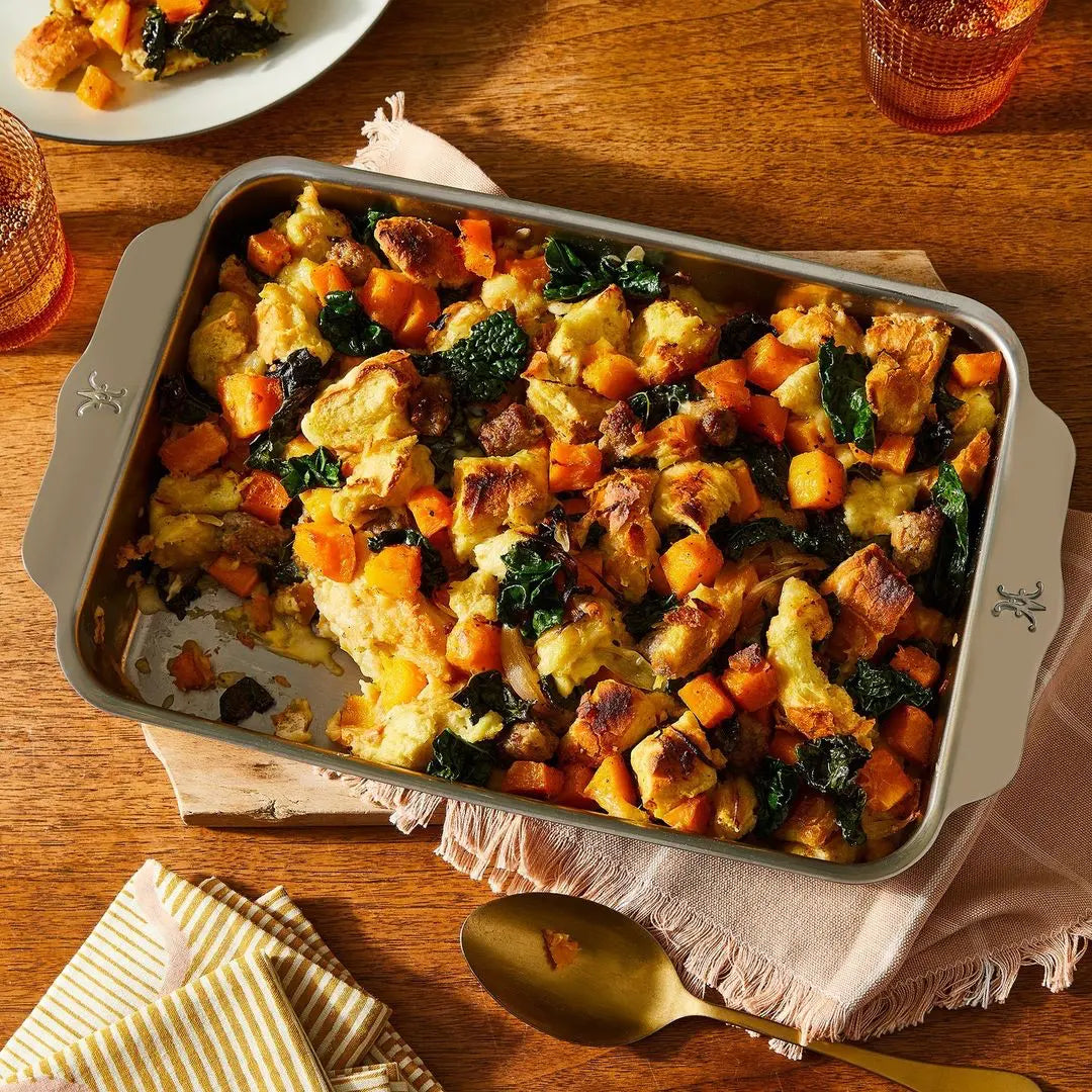 A casserole dish on a wooden table features roasted butternut squash, leafy greens, and sausage, with one serving taken out. The Hestan Provisions OvenBond Tri-ply Small Rectangular Baker ensures even cooking. Nearby are a gold spoon and cloth napkin.