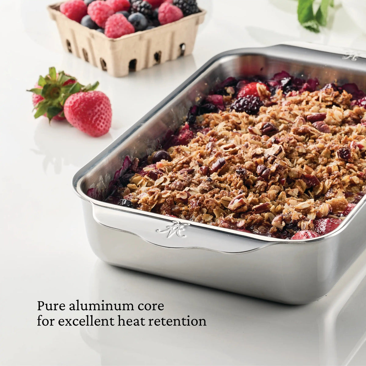 The Hestan Provisions OvenBond Tri-ply Small Rectangular Baker filled with berry crumble showcases its pure aluminum core for excellent heat retention on a white surface, while a nearby container of strawberries adds freshness. Its Clad Stainless Steel construction guarantees durability and even cooking.