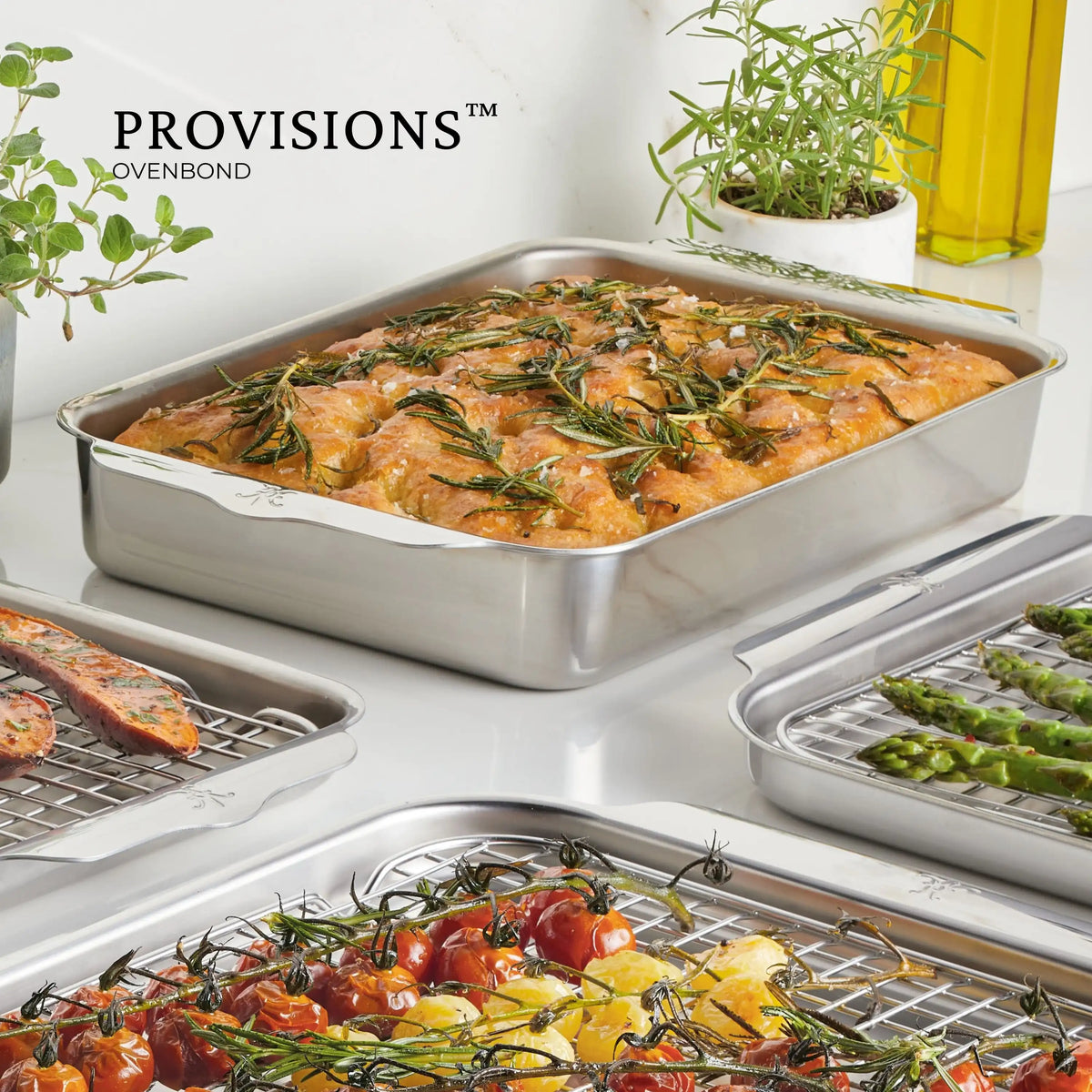 An OvenBond Tri-ply Small Rectangular Baker filled with rosemary-topped focaccia rests on the table. Nearby, trays hold grilled sausages, cherry tomatoes, and asparagus. In the background, a potted plant and a bottle stand beside Hestan Provisions, while its Clad Stainless Steel design ensures its oven safe.