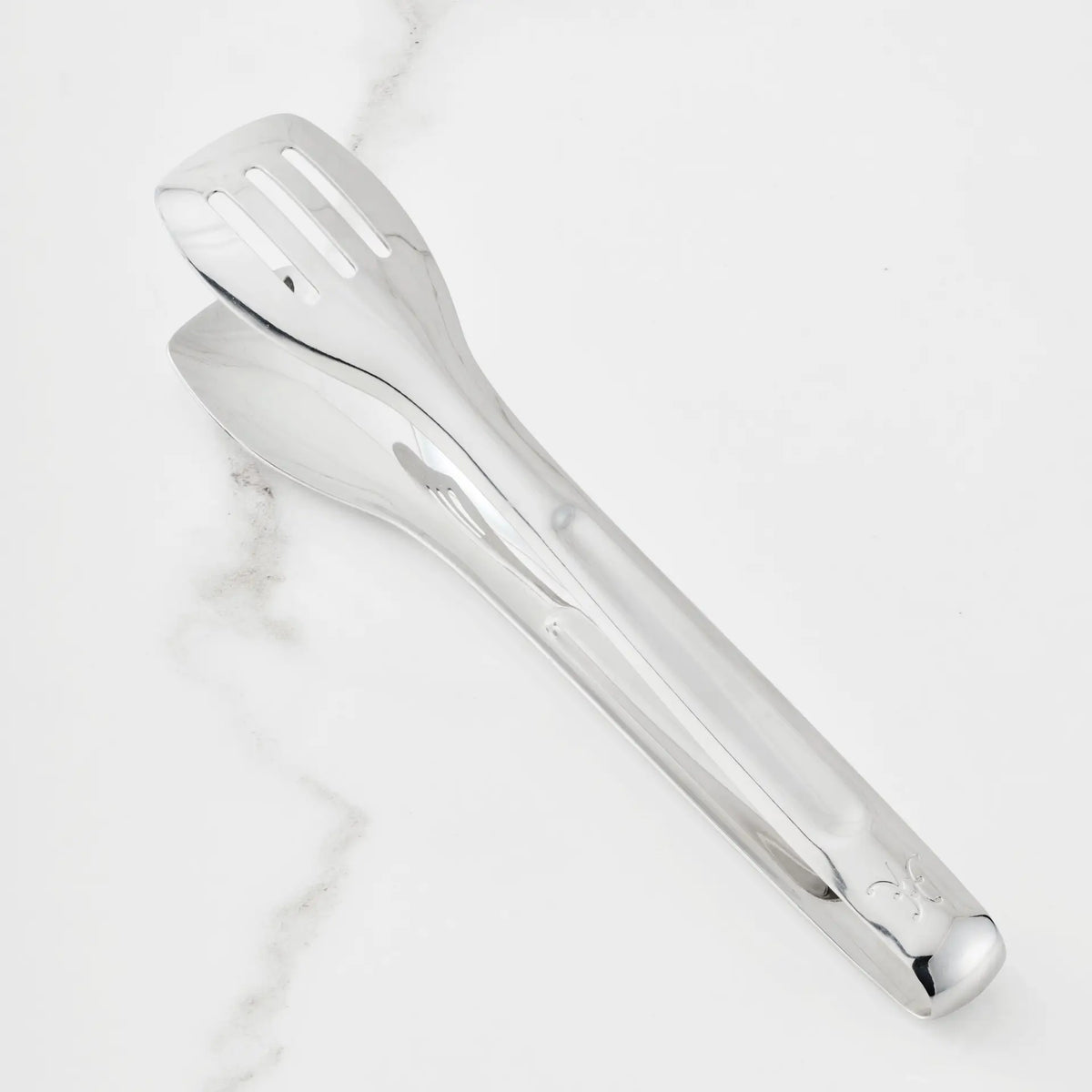 A pair of Hestan Provisions 10-inch Stainless Steel Serving Tongs, with one slotted and one solid side, rest elegantly on a white marble surface featuring subtle gray veining—an essential addition to any ergonomic chef’s tool collection.