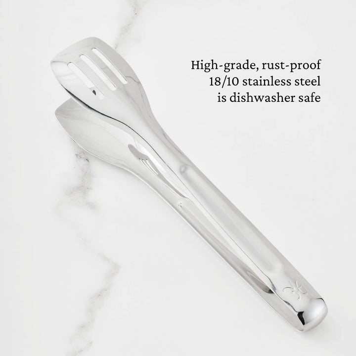 A pair of 10-inch Stainless Steel Serving Tongs by Hestan Provisions with a polished finish rests on a white marble surface. Text beside them reads: Crafted from high-grade, rust-proof 18/10 stainless steel and dishwasher safe—part of the Hestan Provisions collection of ergonomic chef’s serving tools.