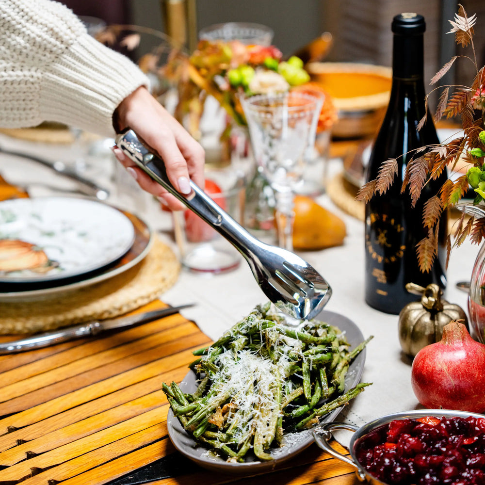 A hand uses 10-inch Stainless Steel Serving Tongs by Hestan Provisions® to lift green beans from an oval platter on a dining table.