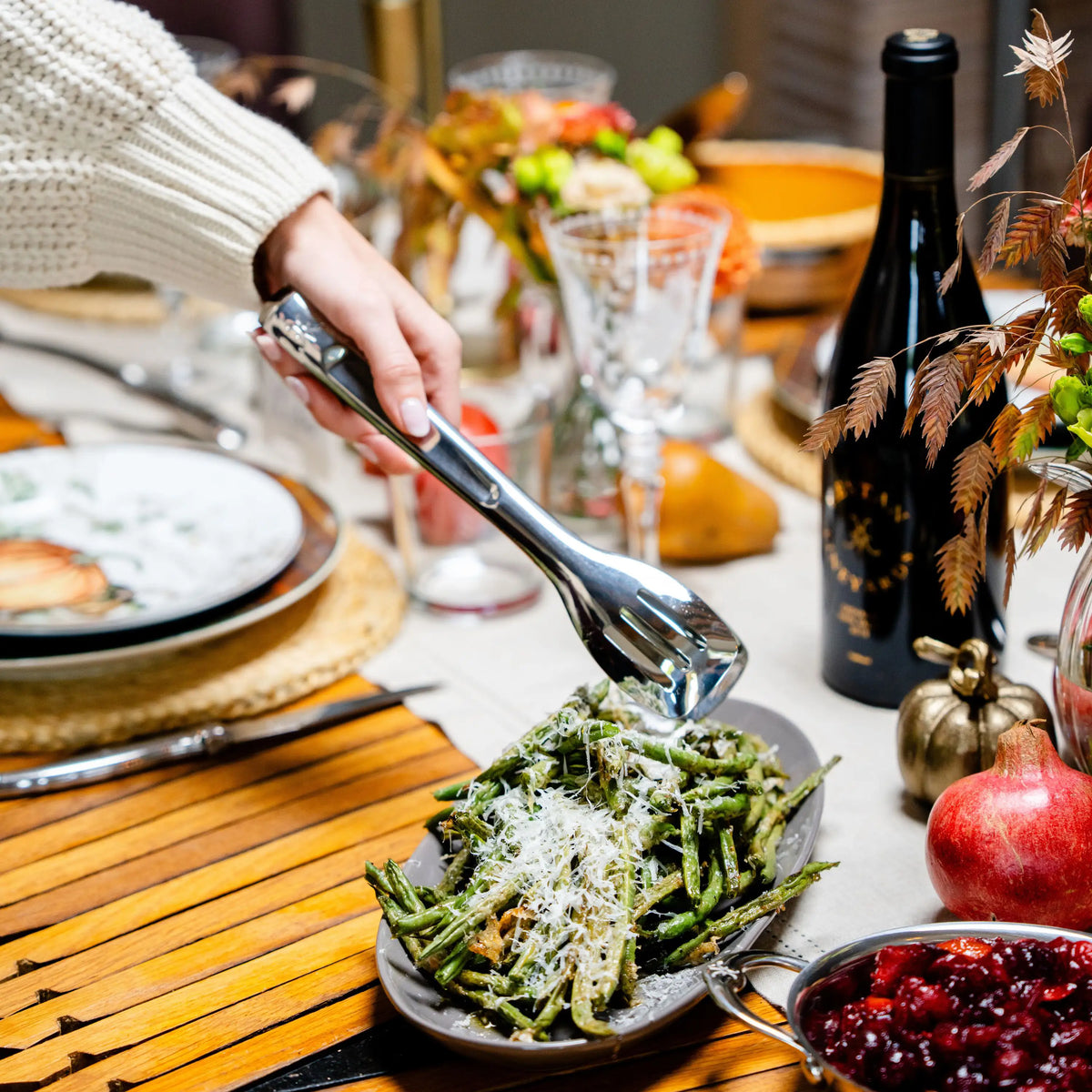 A hand uses 10-inch Stainless Steel Serving Tongs by Hestan Provisions® to lift green beans from an oval platter on a dining table.