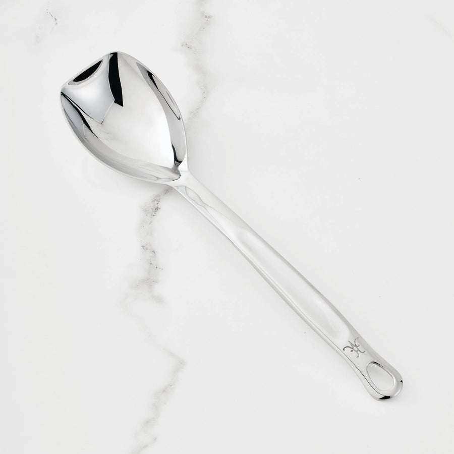 The Hestan Provisions Sauté Spoon with Straight Edge, 13-inch, is a stainless steel serving spoon with a reflective bowl and long handle, designed for ergonomic use—shown here on a white marble surface with subtle gray veining.