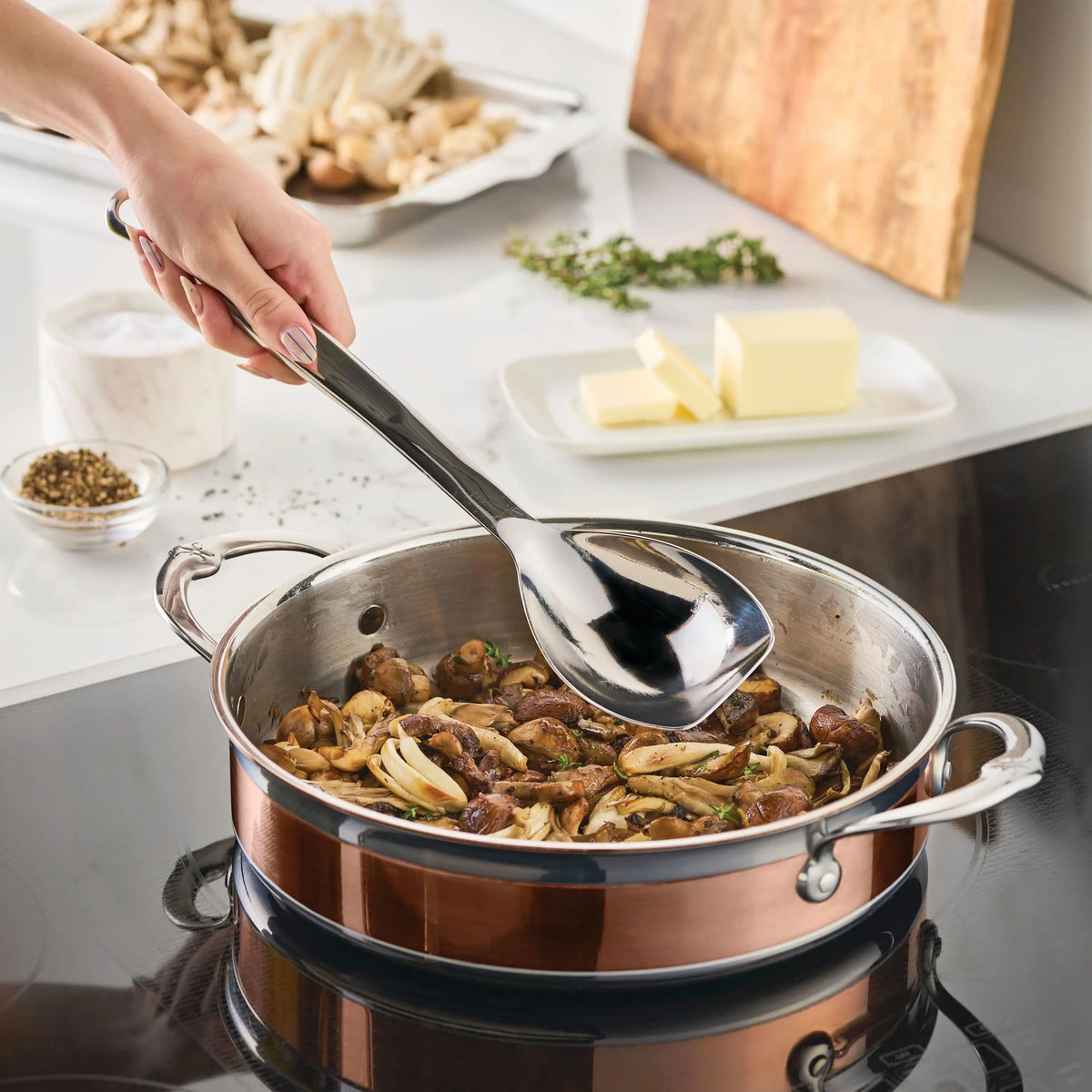 A hand stirs mushrooms in a copper-toned pan on the stove using the 13-inch Sauté Spoon with Straight Edge from Hestan Provisions. Butter, herbs, pepper, and mushrooms are visible on the kitchen counter in the background.