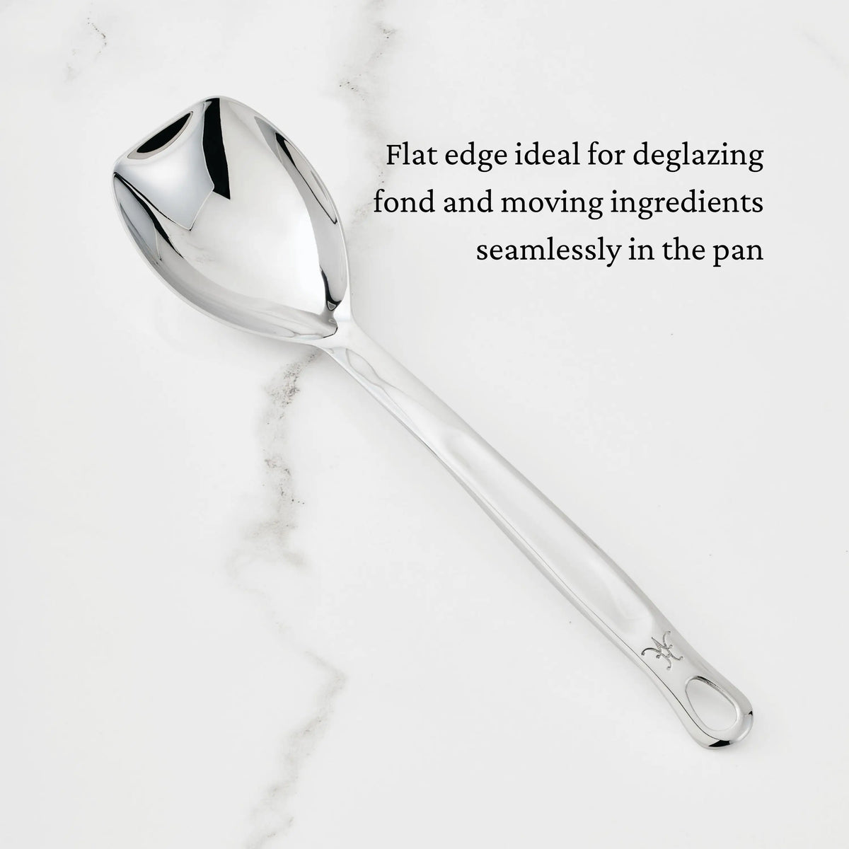 A Hestan Provisions Sauté Spoon with Straight Edge, 13-inch, rests on white marble. Black text reads, “Flat edge ideal for deglazing fond and moving ingredients seamlessly in the pan.”.