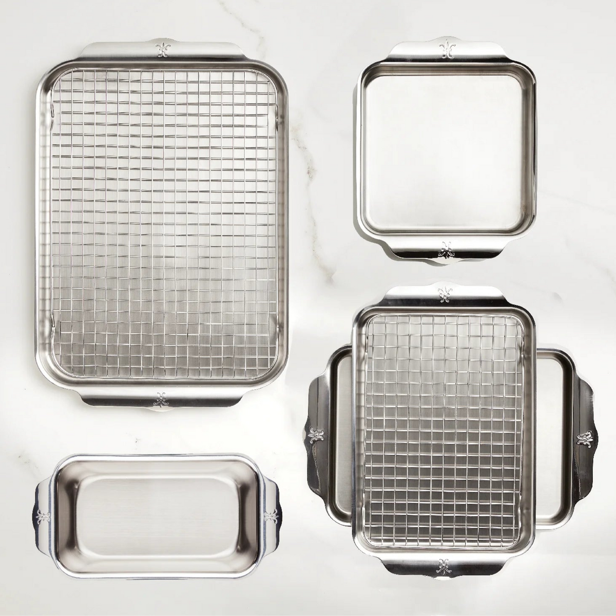 The award-winning Hestan Provisions OvenBond Tri-ply Gourmet Essentials Stainless Steel 7-piece Set is artistically displayed on a white marble surface, featuring a rectangular baking sheet with cooling rack, a smaller pan with rack, another without a rack, and a durable loaf pan.