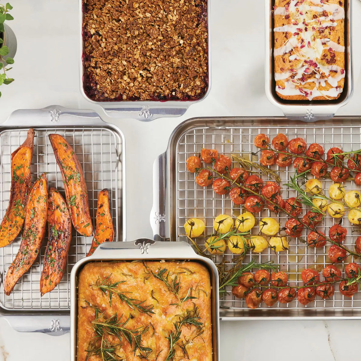 On a white surface, discover an array: berry crumble, glazed loaf cake, herbed sweet potato wedges, and rosemary focaccia—all crafted with the durable OvenBond Tri-ply Gourmet Essentials 7-piece set by Hestan Provisions. Nearby are skewers of cherry tomatoes, pineapple, and red onion on a wire rack.