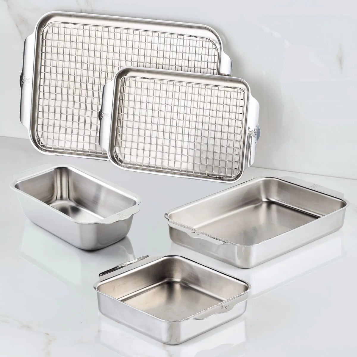 Five pans from the NanoBond Titanium x Tyler Florence 41-Piece Cookware Set, including two with matching wire racks, are arranged on white marble. The pans vary in size and shape, all with straight sides and handles.