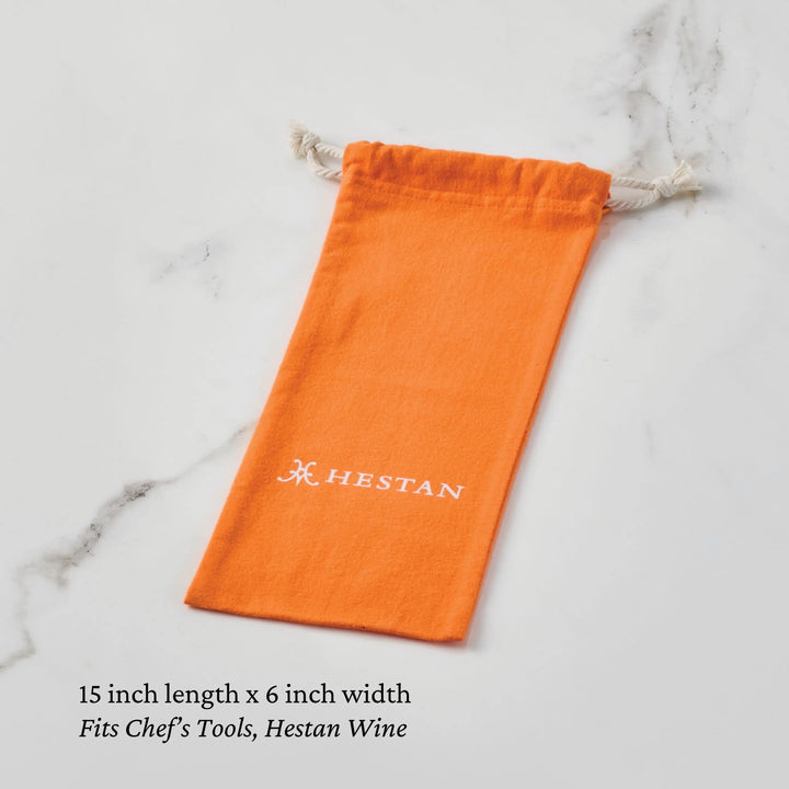An orange Flannel Cookware Storage Bag from Accessories with "HESTAN" in white lies on a light marble surface. Below it reads: 15-inch length x 6-inch width. Ideal for storing cookware, chef’s tools, or protecting Hestan Wine.