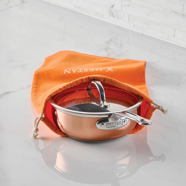 A copper saucepan with a lid sits partially inside an orange Flannel Cookware Storage Bag by Accessories, displayed on a white marble surface.