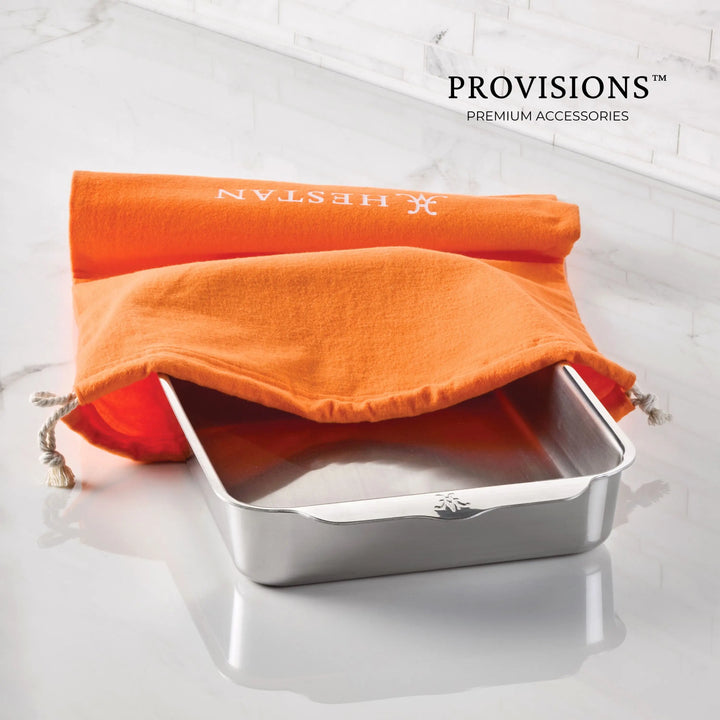 A rectangular stainless steel baking pan is partially covered by an orange Flannel Cookware Storage Bag with white drawstrings. The bag displays the word Accessories. The surface is white marble, and text in the corner reads PROVISIONS PREMIUM ACCESSORIES.