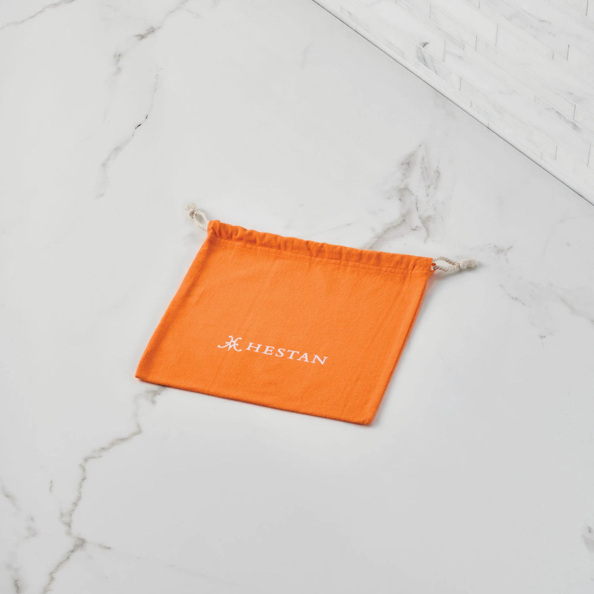 An orange flannel lid storage bag from Accessories, printed with "HESTAN" and a white logo, sits on a veined white marble surface near tile. Ideal for storing cookware or protecting stainless steel lids.