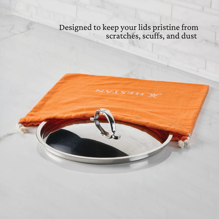 A stainless steel pot lid sits partially inside an Accessories Flannel Lid Storage Bag with white drawstrings on a marble counter. Black text above reads, “Keeps your lids pristine from scratches, scuffs, and dust.”.