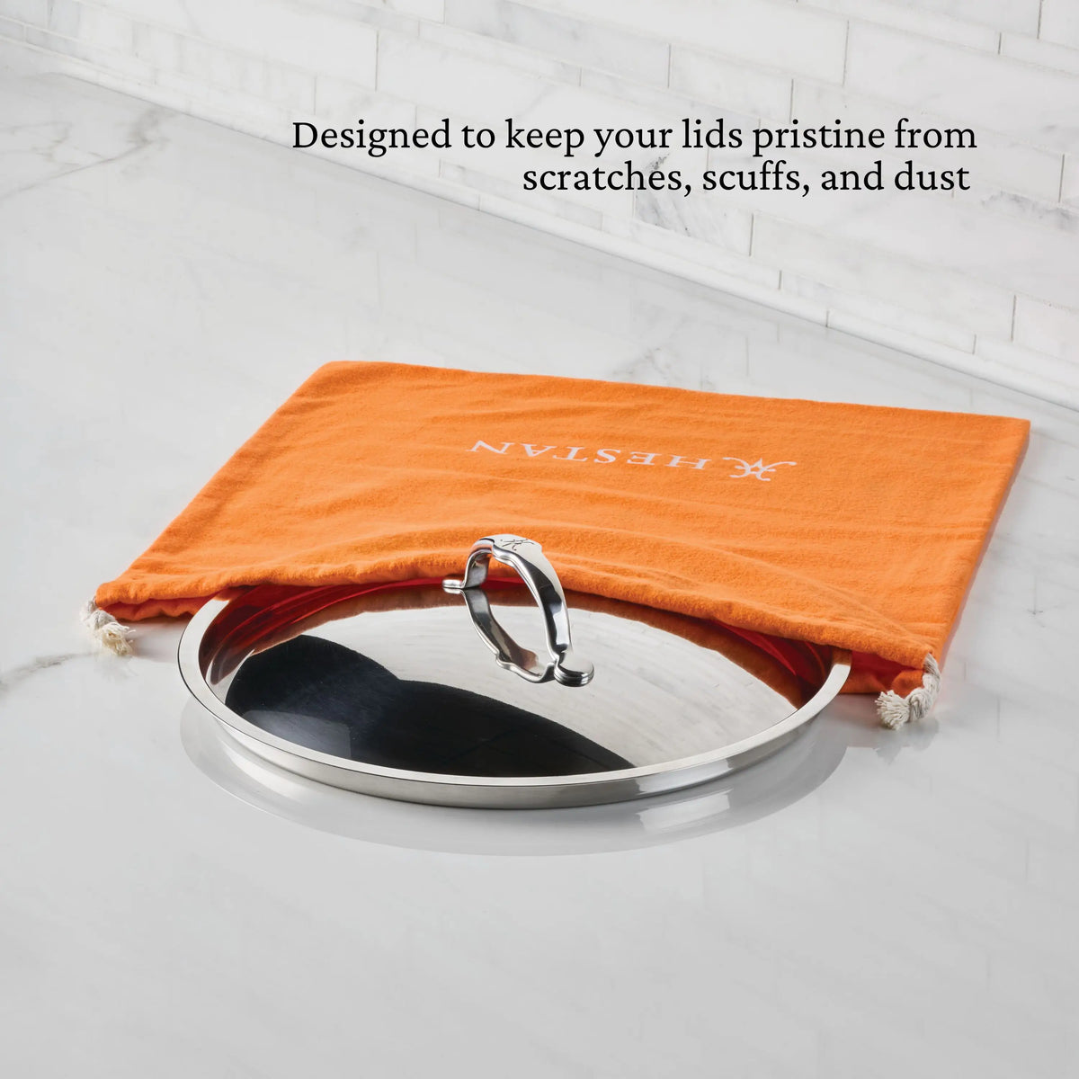 A stainless steel pot lid sits partially inside an Accessories Flannel Lid Storage Bag with white drawstrings on a marble counter. Black text above reads, “Keeps your lids pristine from scratches, scuffs, and dust.”.