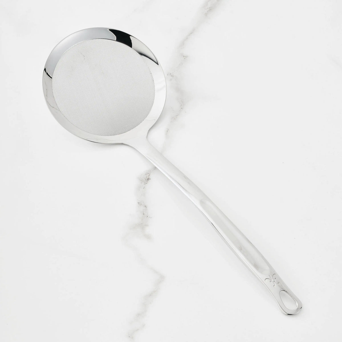 A Hestan Provisions Fine Mesh Skimmer, 16-inch, with a long handle and stainless steel head rests on a white marble surface.