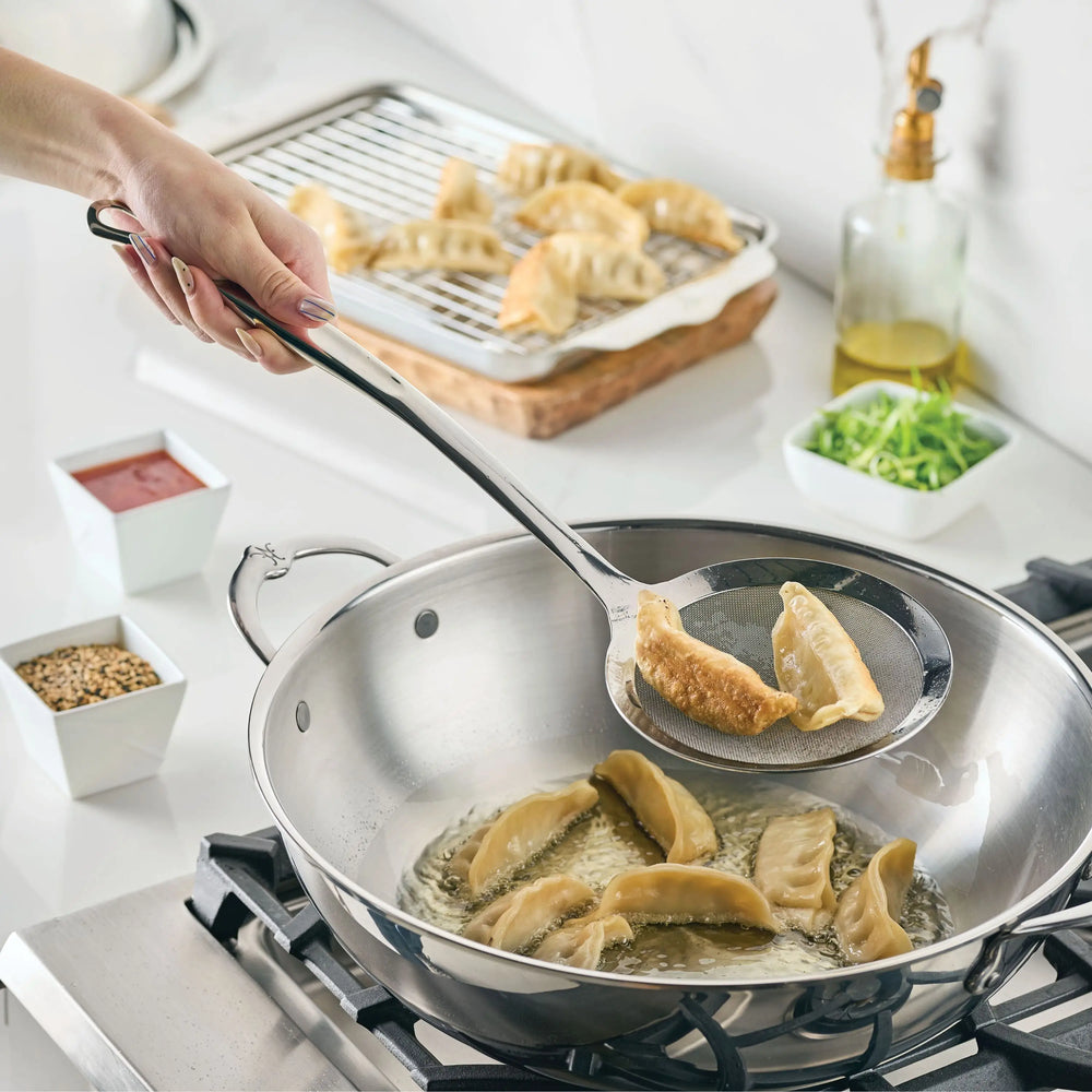 A hand uses the Hestan Provisions Fine Mesh Skimmer, 16-inch, to lift cooked dumplings from hot oil on the stove; more dumplings cool on a rack in the background, surrounded by small bowls of ingredients.