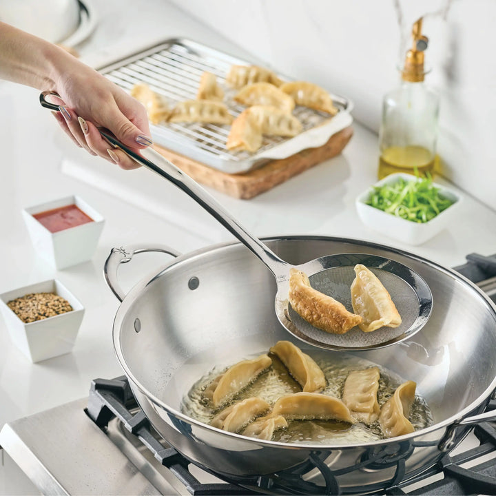 A hand uses the Hestan Provisions Fine Mesh Skimmer, 16-inch, to lift cooked dumplings from hot oil on the stove; more dumplings cool on a rack in the background, surrounded by small bowls of ingredients.