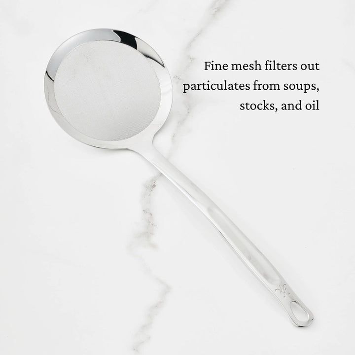 A Hestan Provisions Fine Mesh Skimmer, 16-inch, with a long handle sits on white marble. Text on image: Fine mesh filters particulates from soups, stocks, and oil.