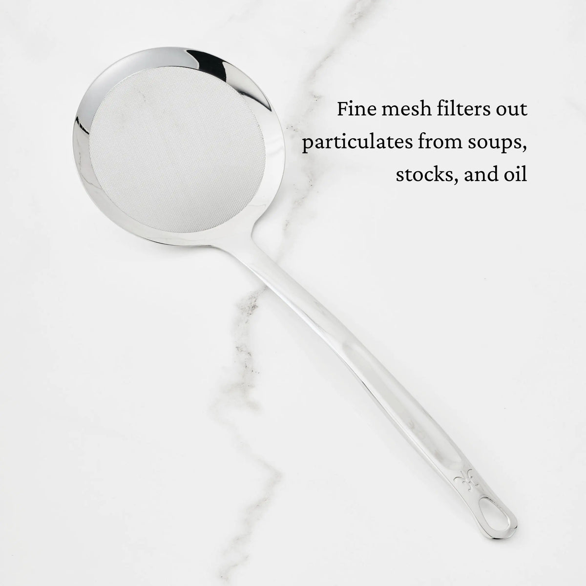 A Hestan Provisions Fine Mesh Skimmer, 16-inch, with a long handle sits on white marble. Text on image: Fine mesh filters particulates from soups, stocks, and oil.