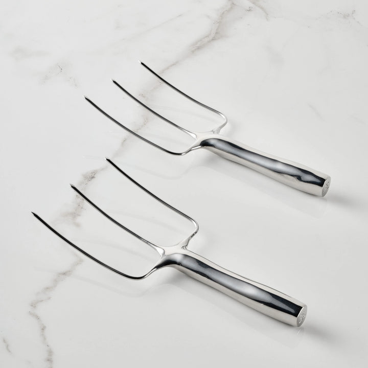 Two Stainless Steel Carving Forks from the Hestan Provisions® set, each featuring three tines, lie parallel on a white marble surface. Made from durable 18/10 stainless steel, their handles are smooth and highly reflective. Arranged with the tines facing upward and to the left, these forks showcase Hestan Provisions®’ dedication to superior quality and design.