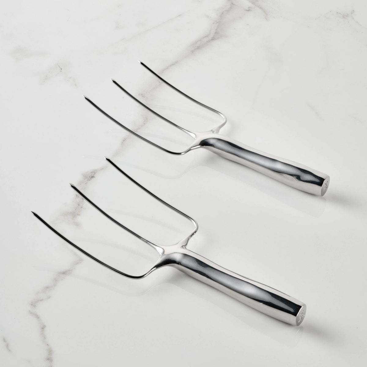 Two Stainless Steel Carving Forks from the Hestan Provisions® set, each featuring three tines, lie parallel on a white marble surface. Made from durable 18/10 stainless steel, their handles are smooth and highly reflective. Arranged with the tines facing upward and to the left, these forks showcase Hestan Provisions®’ dedication to superior quality and design.