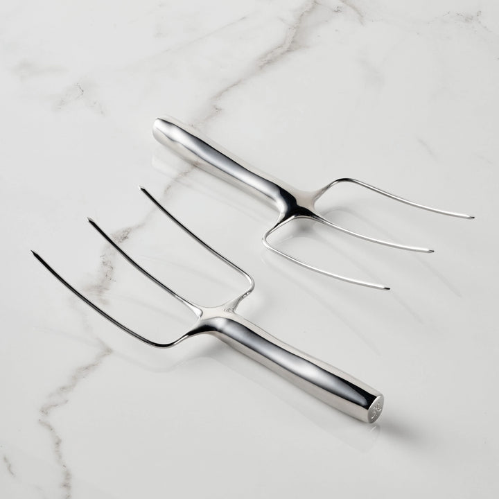 Two Hestan Provisions® Stainless Steel Carving Forks, Set of 2—each featuring three prongs and crafted from durable 18/10 stainless steel—are displayed on a white marble surface, with one fork facing up and the other facing down.