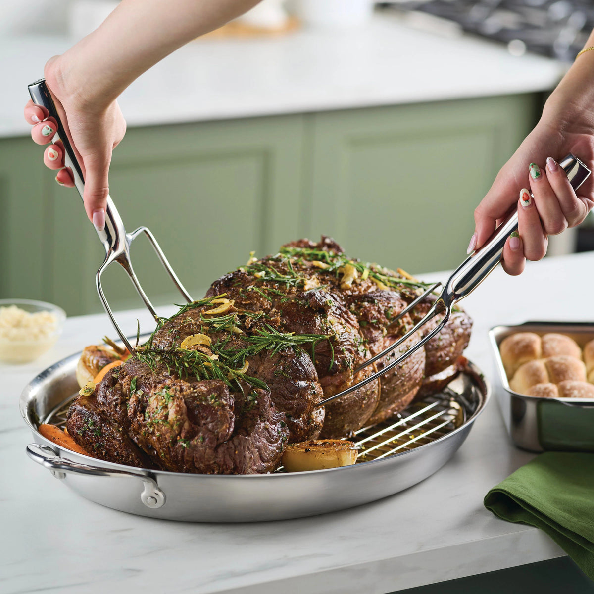 A person uses a Hestan Provisions® Stainless Steel Carving Forks, Set of 2, to lift a seasoned roast from a Hestan Culinary 18/10 stainless steel roasting pan with vegetables. The scene is set on a kitchen counter with rolls and a green napkin nearby.