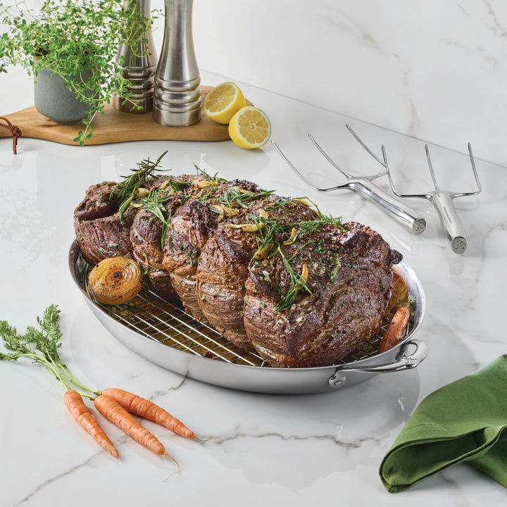 A roasted beef rib roast garnished with herbs rests on a wire rack in a Hestan Culinary 18/10 stainless steel pan, surrounded by carrots and onions. Fresh thyme, lemons, Hestan Provisions® Stainless Steel Carving Forks (Set of 2), salt and pepper shakers, and a green napkin are arranged nearby on a marble countertop.