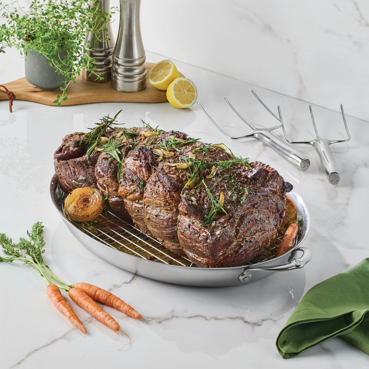 A roasted beef rib roast garnished with herbs rests on a wire rack in a Hestan Culinary 18/10 stainless steel pan, surrounded by carrots and onions. Fresh thyme, lemons, Hestan Provisions® Stainless Steel Carving Forks (Set of 2), salt and pepper shakers, and a green napkin are arranged nearby on a marble countertop.
