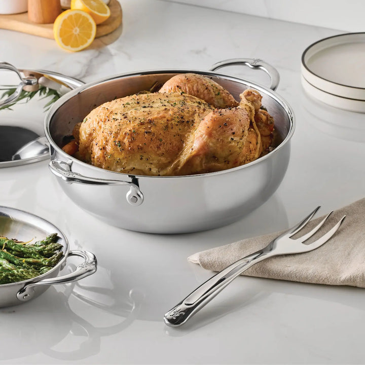 A cooked whole chicken sits in a stainless steel pot on a white countertop. Nearby are a Hestan Provisions 10.5-inch Stainless Steel Serving Fork resting on a napkin, a dish with green beans, stacked plates, and sliced lemons in the background—essential chef’s tools for serving a delicious meal.