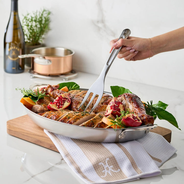 A hand uses a 10.5-inch Stainless Steel Serving Fork by Hestan Provisions® to lift a slice of roasted chicken from a platter garnished with pomegranate, orange slices, and herbs.