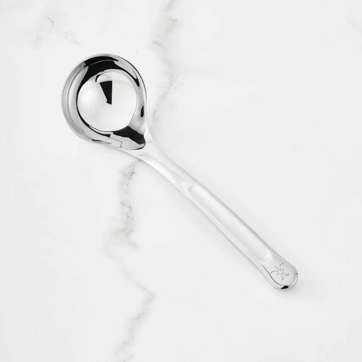 A 2.4-oz Stainless Steel Serving Ladle from Hestan Provisions, distinguished by a decorative detail near the end of its handle, rests on a white marble surface with subtle gray veining.