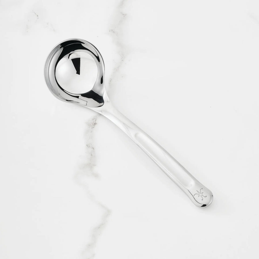 A 2.4-oz Stainless Steel Serving Ladle from Hestan Provisions, distinguished by a decorative detail near the end of its handle, rests on a white marble surface with subtle gray veining.