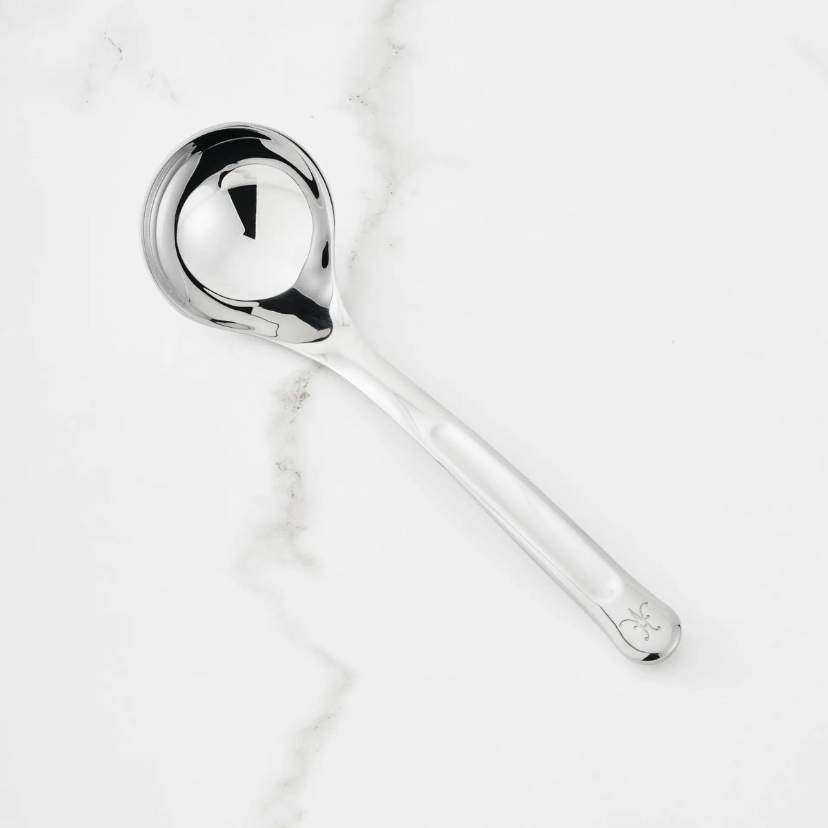 A 2.4-oz Stainless Steel Serving Ladle from Hestan Provisions, distinguished by a decorative detail near the end of its handle, rests on a white marble surface with subtle gray veining.